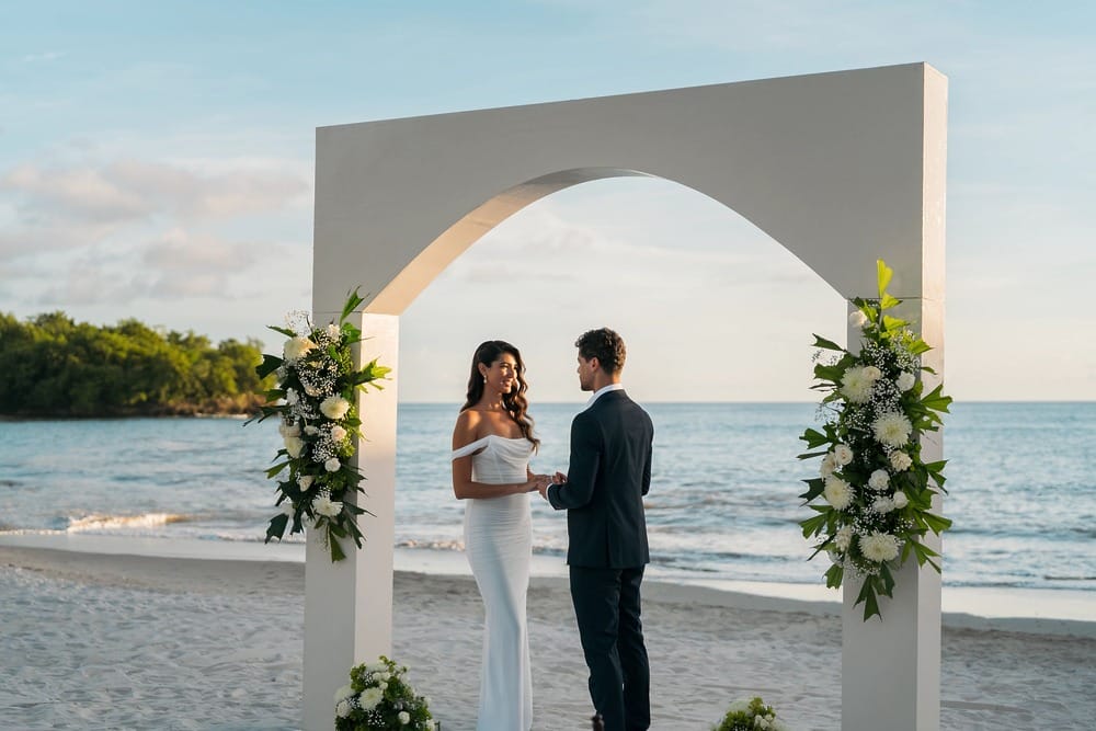 A couple at their destination wedding stands under a decorated arch on the beach.