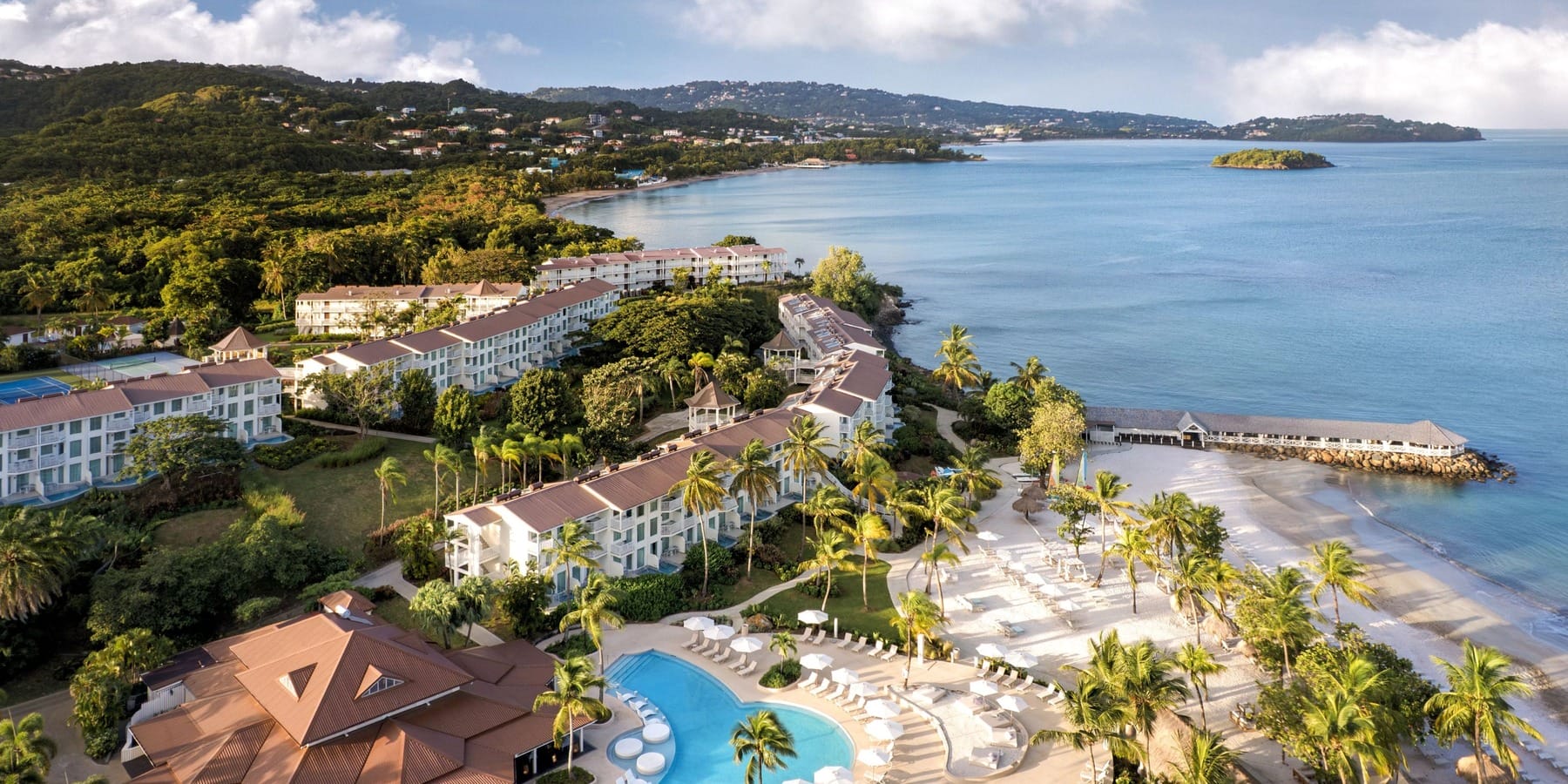 Aerial view of a beachfront destination wedding resort with pools, palm trees, and nearby hills.