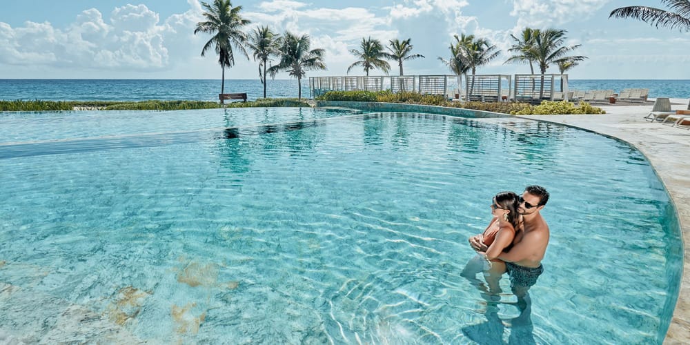 A couple enjoys a resort pool at a tropical destination wedding with ocean and palm trees behind.