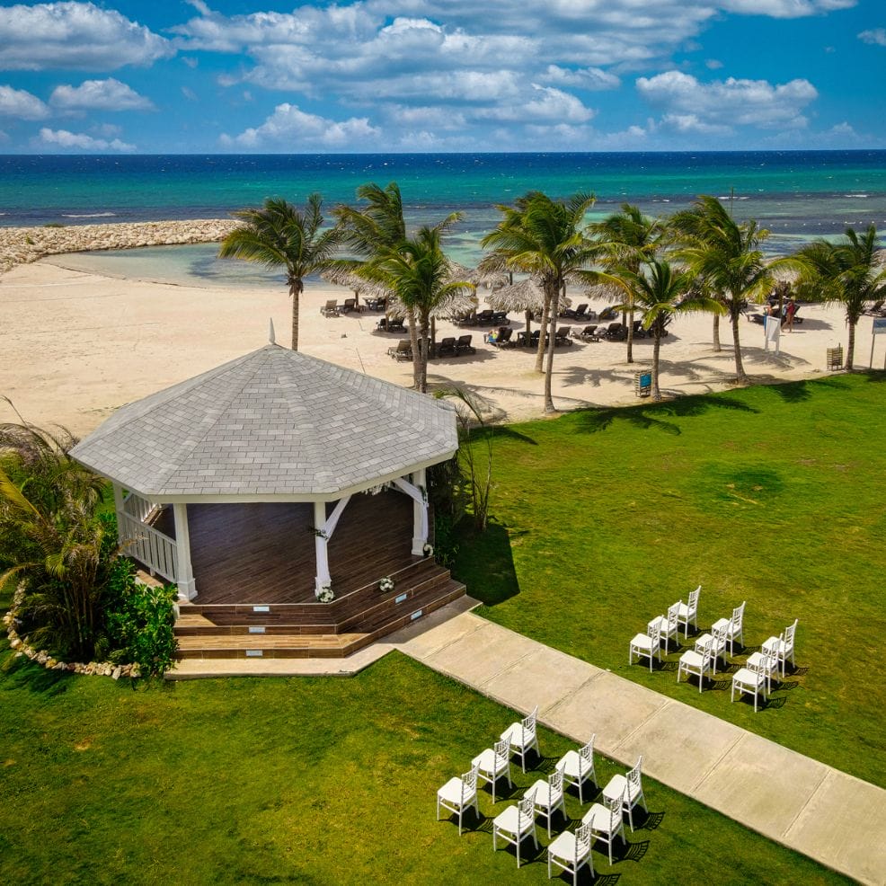 White gazebo with chairs set up for a destination wedding on a lawn by the beach and palm trees.