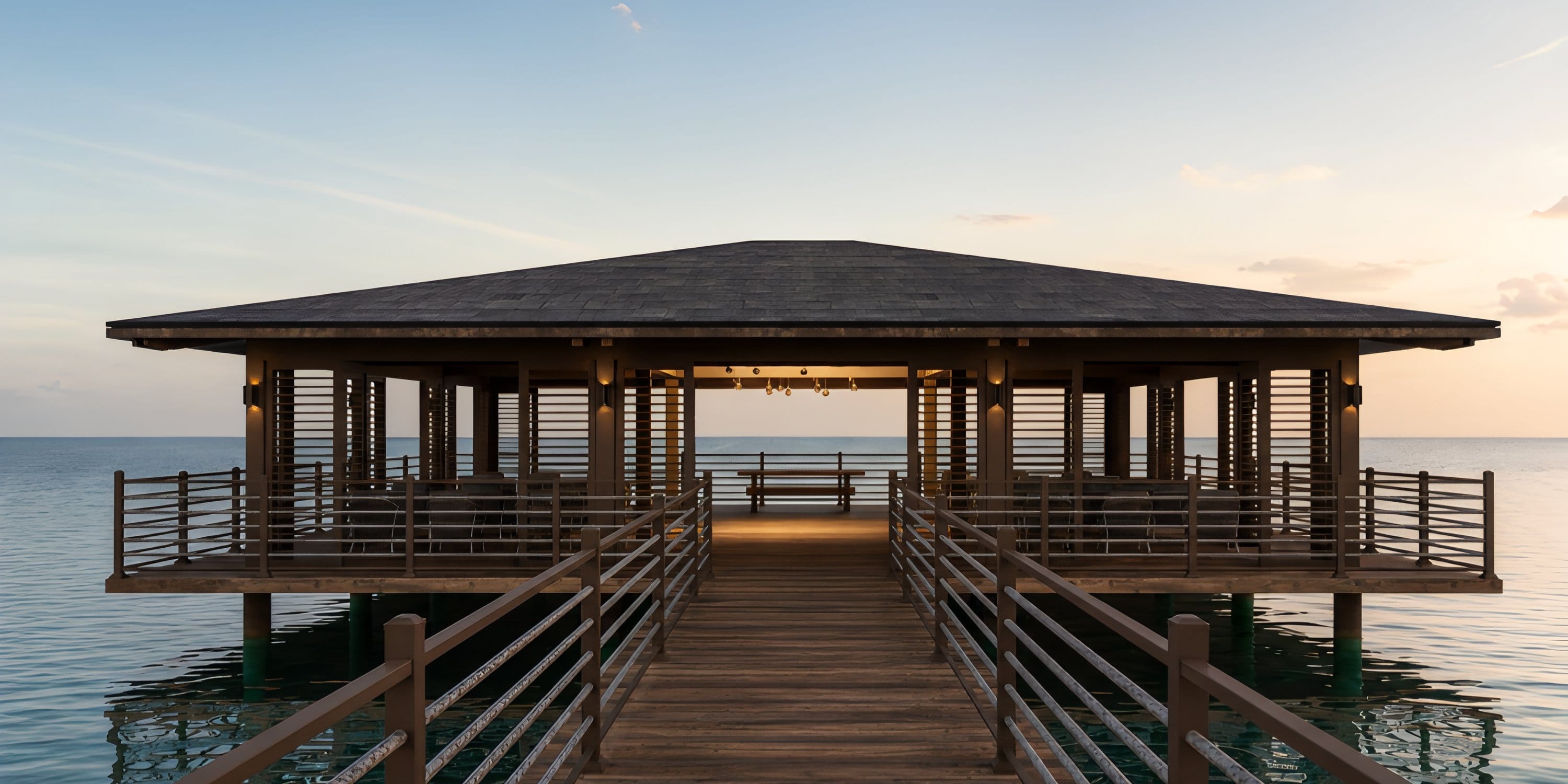 A wooden pavilion on stilts extends over calm water at sunset, perfect for a destination wedding.