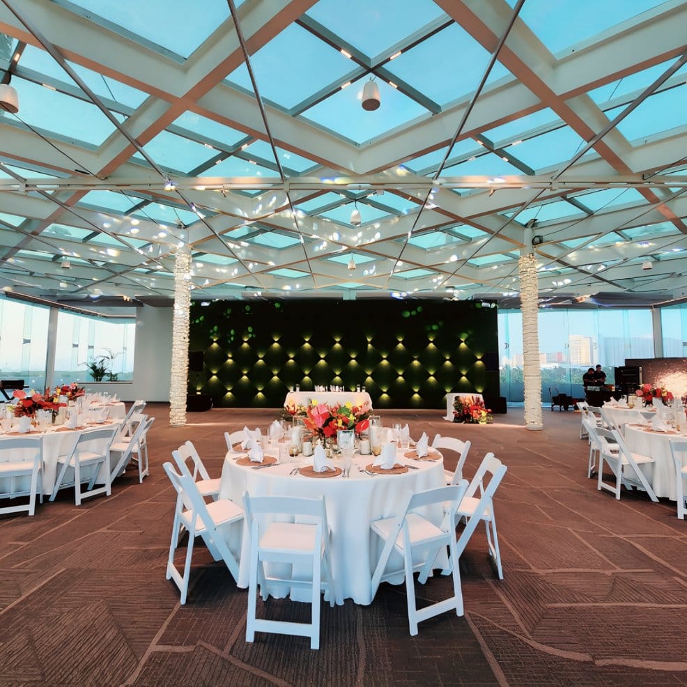 Round tables set for a wedding reception under a glass grid ceiling.