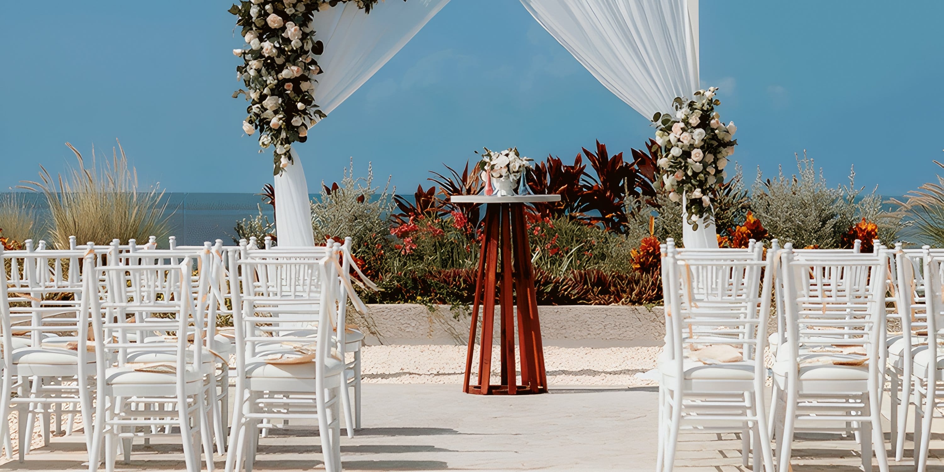 White wedding chairs facing a flower-decorated altar by the ocean at Dreams Playa Mujeres.