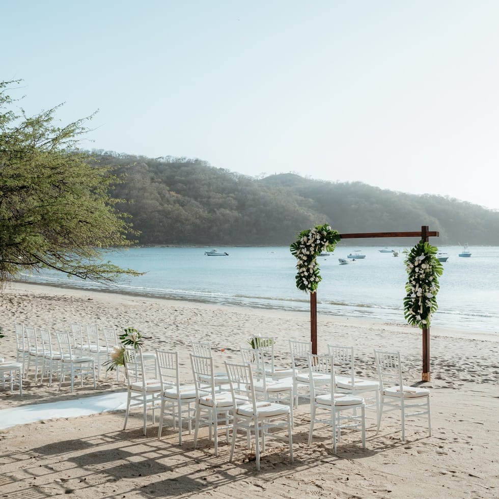 Destination wedding arch with white flowers and chairs set up on a sandy beach.