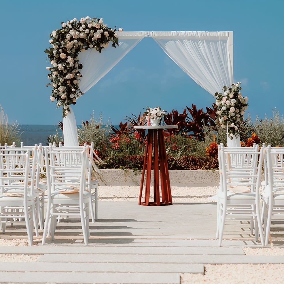 White chairs for a destination wedding at an altar with roses near the ocean.