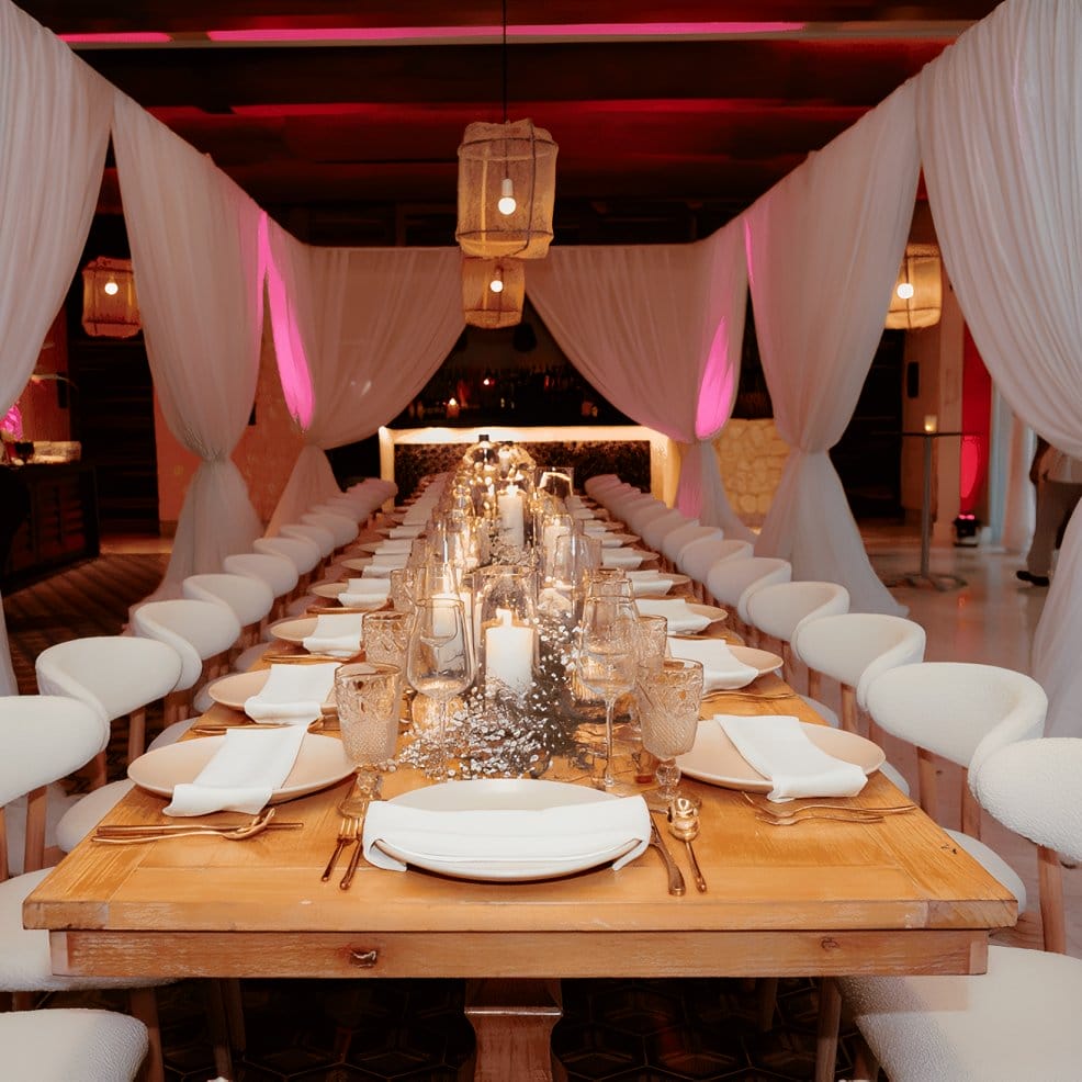 Long wooden table set for a destination wedding, lined with candles and white draped curtains.